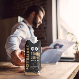 Person holding a Joyburst Protein Coffee Dark Roast package on a table with a blurred background