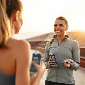 Two women in workout clothes, at sunset, one holding a protein shake container.